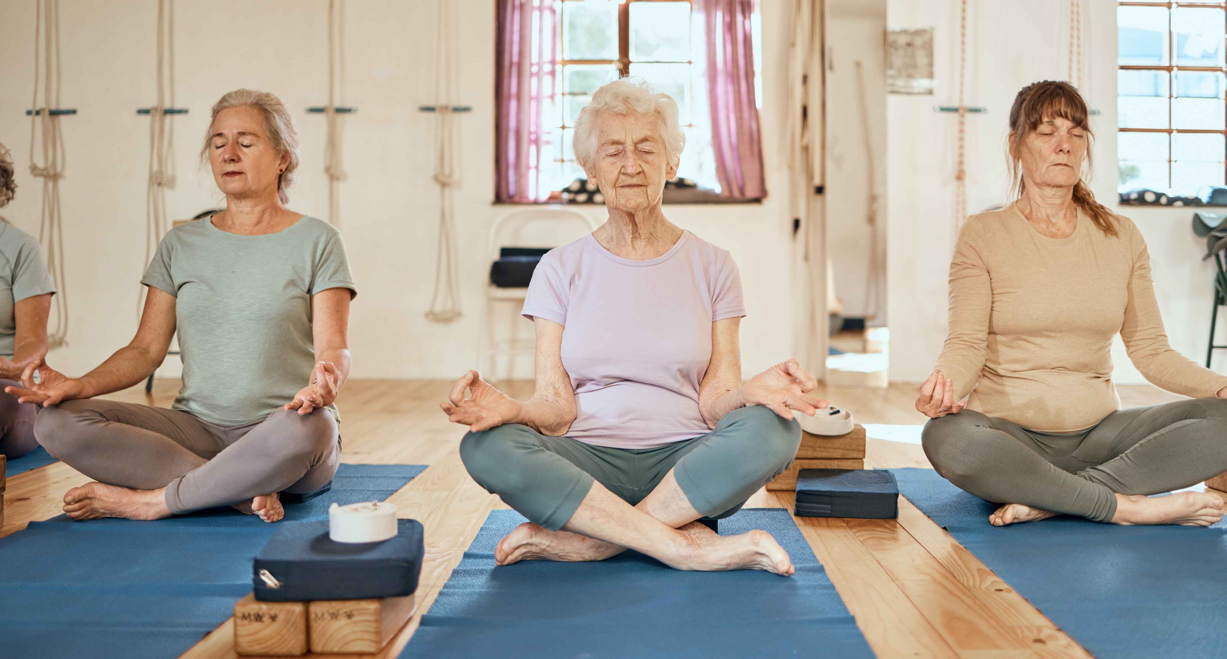 Group of senior women sitting cross-legged on yoga mats with eyes closed, practicing meditation in a calm wellness studio.